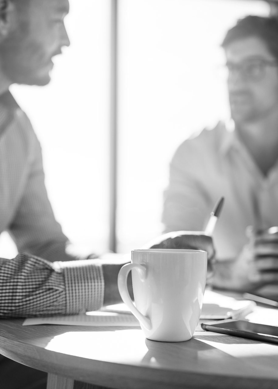 Business executives discussing work at office. Closeup of coffee cup with blurred image of two businessmen sitting on table.